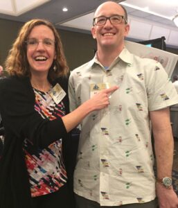 This is a photograph from a Dysphagia Research Society meeting in San Diego in 2019 of Karen Sheffler pointing to John Holahan's newly designed IDDSI button down/collared shirt with IDDSI framework and IDDSI syringes on a beige background. You can purchase one at the link in the bulleted section above this photograph. 